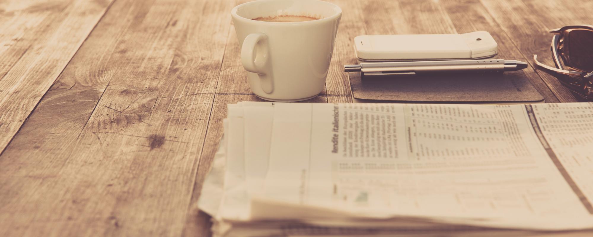 folded newspaper on a desk with a coffee cup, phone and sunglasses